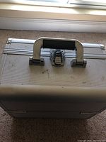 Top view of small worn silver makeup case showing textured surface, central lock, and black handle.