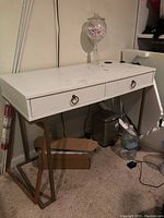Photo showing white writing desk with two drawers, gold accent ring handle hardware, and angular gold metal legs.