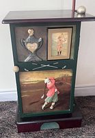 Front view of golf putting cabinet showing the decorative front door with 3 golf-themed panels: 'First Place' trophy, framed vintage golfer print, painted golfer in swing, and visible top putting area with golf ball and putter.