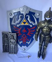 Photo showing the Legend of Zelda shield with blue, silver, red, and gold paint, two metal wall plaques with knight relief, small metal sword, and metal knight statue in bronze armor.