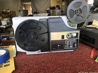 Side view of Bell and Howell Maxa 8mm film projector showing reel holder, control panel with switches and knobs, and overall mechanical layout