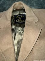 Close-up photo of the camel color cashmere coat showing the label '100% Pure Mongolian Cashmere Hughes & Hatcher Collection' inside the coat lining.