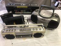 Three radio cassette tape players, including Sharp, Sony, and Oritron models, displayed together on a white surface with power cords visible.