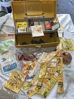 Yellow plastic fishing tackle box opened with various fishing accessories inside including hooks, sinkers, leader connections, and bait lines mostly in original packaging laid out in and around the box.