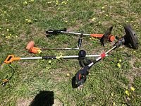 Four assorted weed trimmers/eaters laid on the grass, showing straight shafts, handles, and cutting heads. Various orange and black colors visible.