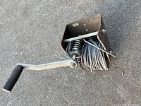 Winch viewed from the side showing the crank handle and coiled steel cable on drum with thick metal mounting bracket and spring