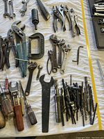 Photo showing various hand tools like pliers, wrenches, clamps, screwdrivers, and drill bits arranged on a white cloth.