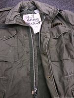 Close-up photo of the olive green vintage US Army jacket showing the zipper, button closure, right chest U.S. Army embroidery, and name tag on left chest pocket.