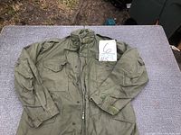Full view of military field jacket laid flat on gray surface showing front pockets, snap buttons, and collar area.