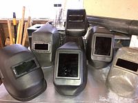 Six welding masks of various styles and sizes with attached lenses placed on a table along with welding rods and containers