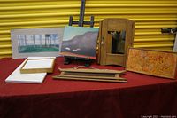 Full lot on red cloth surface with artworks, cabinet, easel and framing materials visible