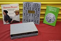 Five art and design books on a red surface against a yellow background: Forever Picasso, Art Deco, Ceramic Design, and two unnamed white-covered volumes.