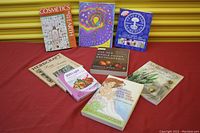 Full view of 8 books arranged on a red surface with golden yellow background. Titles visible: Cosmetics From The Earth, The Magical Sun, Neal's Yard Remedies, Herbcraft, The New Whole Foods Encyclopedia, 200 Light, Original Herbal Magic, Alfreda's Herbs.