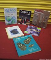 Six art books arranged on red cloth with yellow shutter background, showing titles related to glass, ceramics, woodworking, wallpaper, and craft traditions.