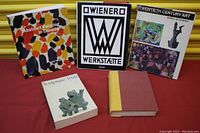Five art books arranged on a red surface with yellow background, visible titles and spines. Includes colorful abstract painting book, Wiener Werkstätte, Twentieth Century Art, Sculpture Inuit, and a red-yellow cloth bound book.