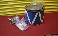 Photo of vintage circus drum container with lidded top and several bundles of candles and ribbons placed beside it on red fabric.