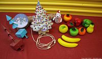 Full lot with ceramic Christmas tree, glass fruit, bear figurine, wooden tree sculptures, and snowman music box arranged on red fabric.