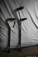 Photo shows two canes on a gray fabric surface, one standard cane with a curved handle and wrist strap, the other a forearm crutch with adjustable height and black grip handle.