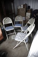 Four white metal folding chairs with cushioned seats arranged in a storage or garage area with boxes and other items in background.