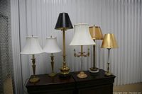 Photo showing six table lamps with various heights, brass and gold bases, white, black, cream, and gold shades on a wooden surface in front of a corrugated metal wall.