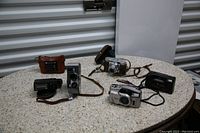 Photo showing six assorted cameras of various types on a round table, including vintage and modern models, leather cases and straps visible.