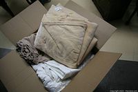 Open cardboard box with assorted queen size bedding showing beige blanket folded on top and several sheets underneath in white, cream, and beige colors.