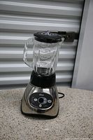 Oster blender with clear glass jar and black lid on a countertop with metal shutter background