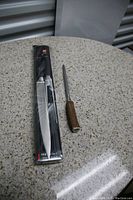 Photo of a carving tool with a wooden handle next to a packaged Zwilling J.A. Henckels Twin knife sharpener on a round textured table.