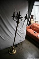 Full view of the brass floor lamp with five candle-style electric arms, standing against a white backdrop in a storage space.