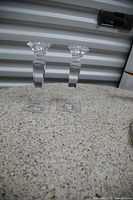 Photo shows two clear glass candle sticks with square bases and faceted tops placed on a speckled countertop against a corrugated metal background.