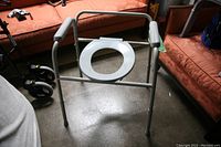 Photo of the raised over toilet seat frame with handles and white open-front plastic seat, showing overall condition and dimensions relative to surrounding furniture.