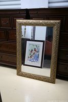 Large rectangular mirror leaning against a dark wood dresser, showing its decorative leaf patterned gold-toned frame and clear reflective surface.