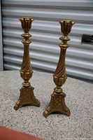 Pair of gold-colored wooden candle sticks with ornate triangular bases and twisted stems shown standing on concrete surface.