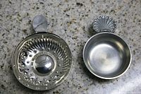 Two silver holders placed on a granite countertop; one holder has a scallop shell-shaped handle and a smooth rounded bowl, the other has a flat handle and a bowl with raised center and dimples.