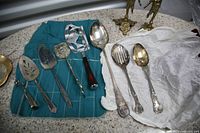 Photo of eight silverware utensils including serving spoons, forks, and scoops, some tarnished, arranged on cloth and tray.