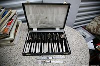 Open black fitted case with ivory satin lining showing six knives, six forks and a sugar spoon