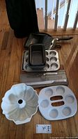 Photo of various metal baking pans including loaf pans, a muffin pan, a cake pan, skillet, a white bundt cake pan and a white microwave egg/muffin pan with multiple compartments, displayed on hardwood floor