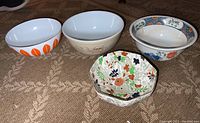 Four bowls displayed on carpet: one octagonal floral bowl, one white Pyrex bowl with orange leaf pattern, one white bowl with muted scenery design, and one round Royal Cauldon bowl with floral rim.