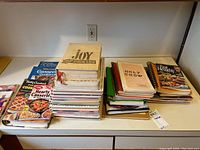 Stacks of assorted cooking magazines, spiral-bound cookbooks, and hard-sided cookbooks on a kitchen counter. Titles include 'Joy of Cooking' and 'Holy Chow.'