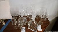Various clear glassware items including two glass decanters and multiple stemmed wine glasses on wooden surface.