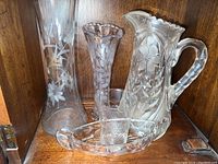 Three vases, glass pitcher, and nut dish on wooden shelf showing etched floral patterns clearly.