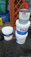 Four containers of paint and primer grouped on the ground outside: two Kilz 2-gallon buckets of All-Purpose 2 primer, one Kilz 1-gallon can of Pro-X Interior Primer PVA, one Behr 2-gallon bucket of ceiling paint.