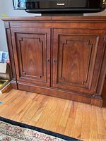 Front view of sidebar cabinet with two paneled doors, warm brown finish, placed on wood floor under a Panasonic TV.