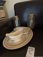 Lenox fine china gravy boat, bowls and platter arranged on a dark surface with two gold-toned rimmed glasses visible beside them.