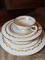 Stacked white Lenox china pieces with gold leaf pattern and gold trim including cups, plates, and bowls.