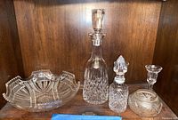 Set of pressed glass items including two bowls, two glass bottles with stoppers, and two candlesticks, displayed on wooden shelf