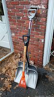 Three tools resting against a brick wall: two snow shovels with aluminum scoops and one orange ice chipper with wooden handle.