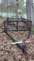 Full view of the weathered metal utility trailer frame set among fallen leaves. Frame is missing a bed and tires appear soft. Reflector poles lay across the front section.