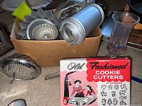Photo of box filled with assorted vintage kitchen utensils including metal molds, shaker, perforated tool, and plastic glass.