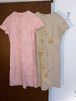 Front view of two vintage linen dresses hanging side by side, showing floral embroidery details and overall style.